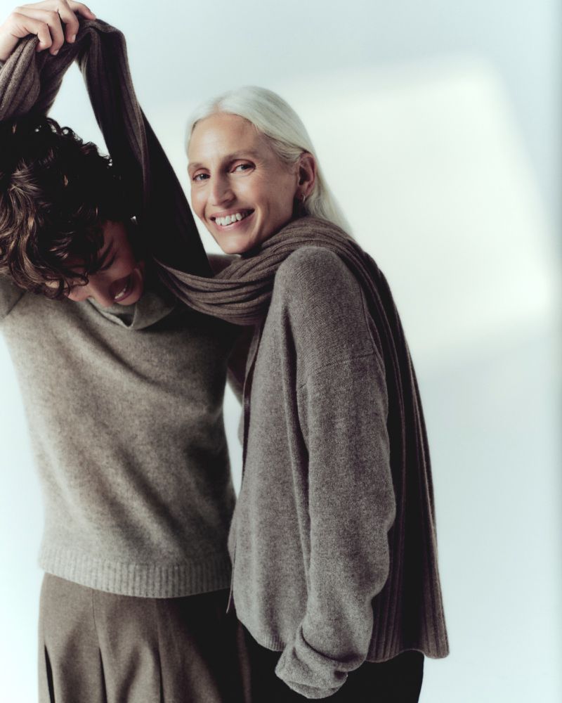 Two people, one older woman with silver hair and a younger person, stand closely together in matching taupe knitwear, smiling in a photo studio.