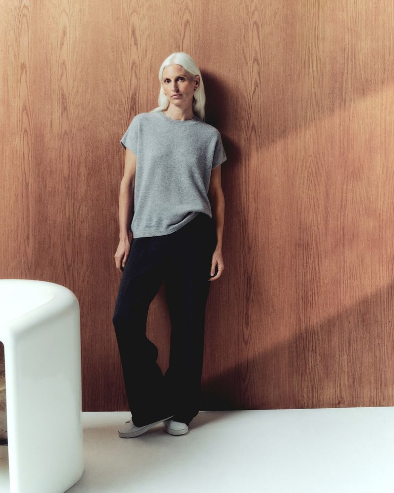 Full-length shot of a woman with platinum blonde hair in a grey shirt and dark pants posing indoors in front of a wood-panelled wall.