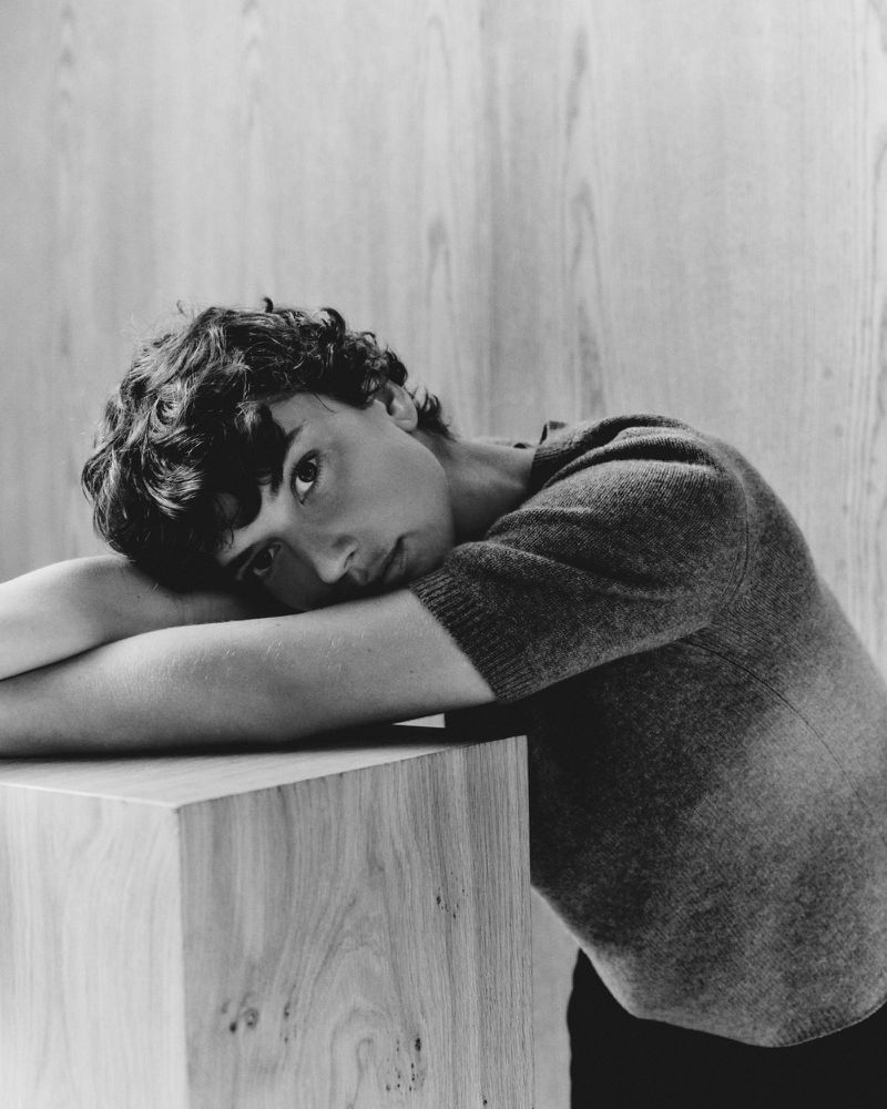 Monochrome portrait of a young adult male with curly dark hair resting his head on a light-colored wooden cube, indoor setting with neutral background.
