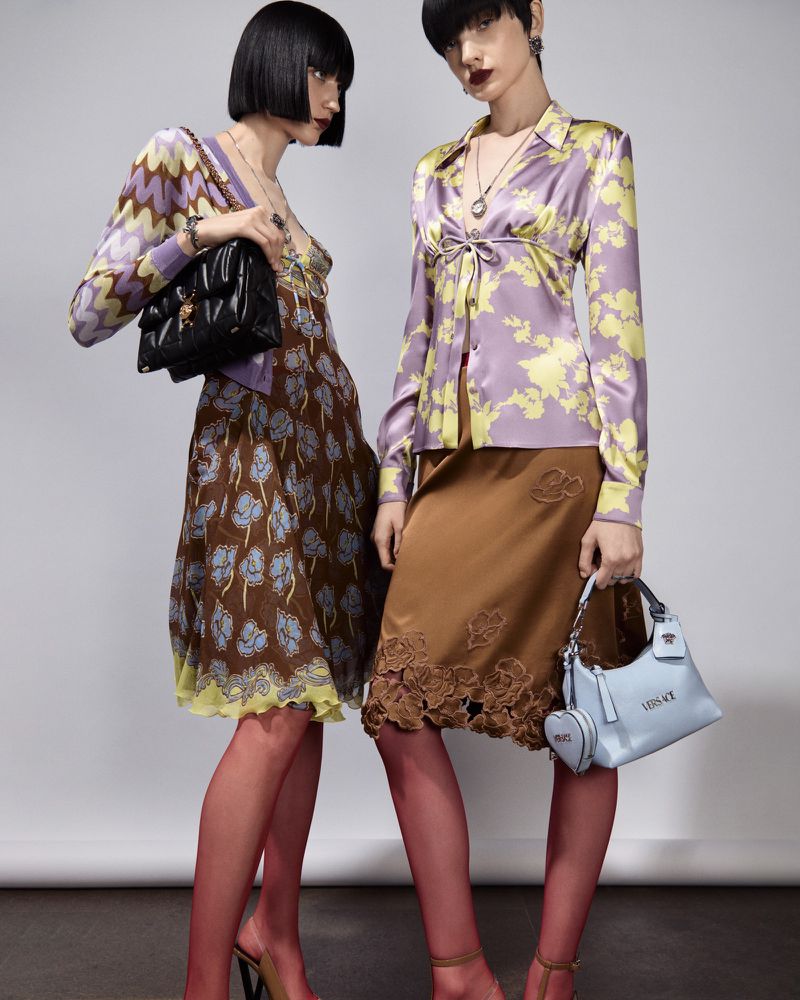 Two women pose in floral satin outfits with vintage purses against a neutral background, showcasing fashion and style.