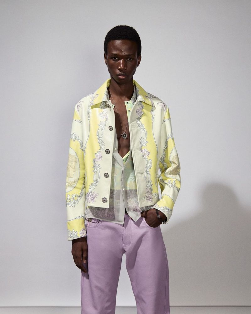 Male model poses indoors wearing a patterned jacket, button-down shirt and lilac pants. The shot is a medium shot on a white background.