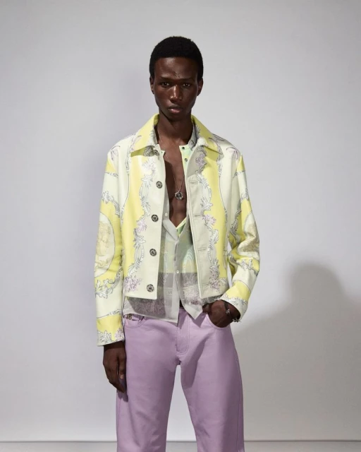 Male model poses indoors wearing a patterned jacket, button-down shirt and lilac pants. The shot is a medium shot on a white background.