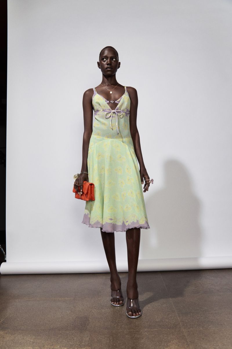 Adult female fashion model poses in light-green floral dress with orange purse in a studio setting. Full-length shot, white backdrop.