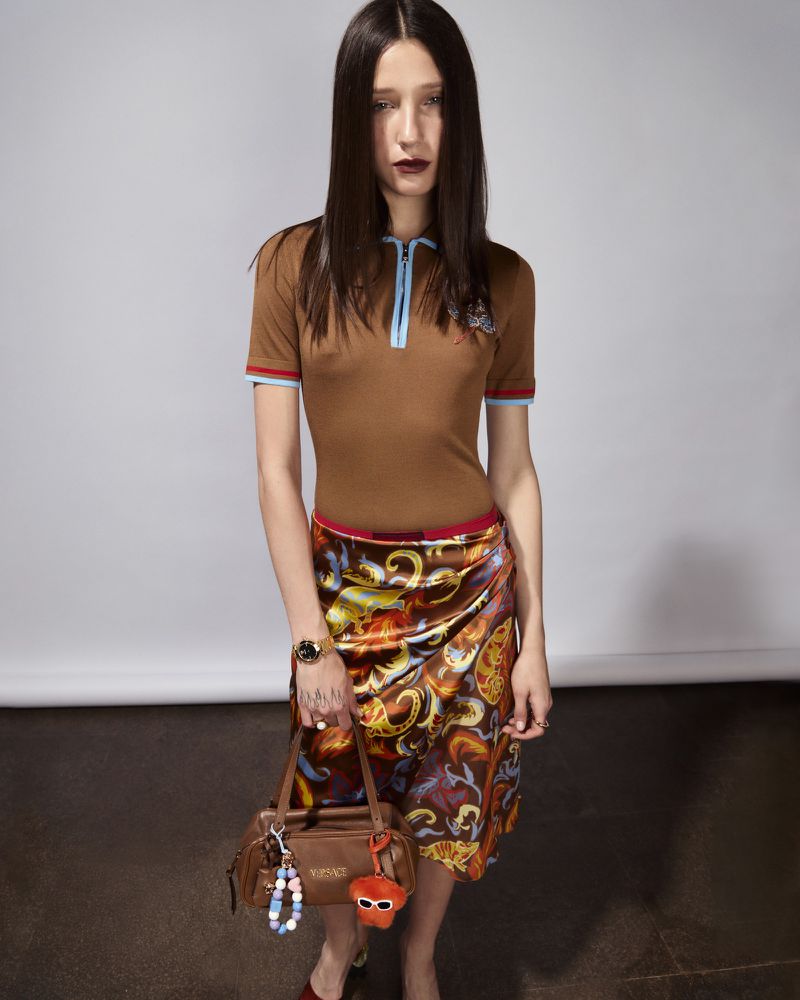 Woman in brown top and patterned skirt holding brown handbag with beaded keychain, standing in a photo studio with a neutral backdrop.