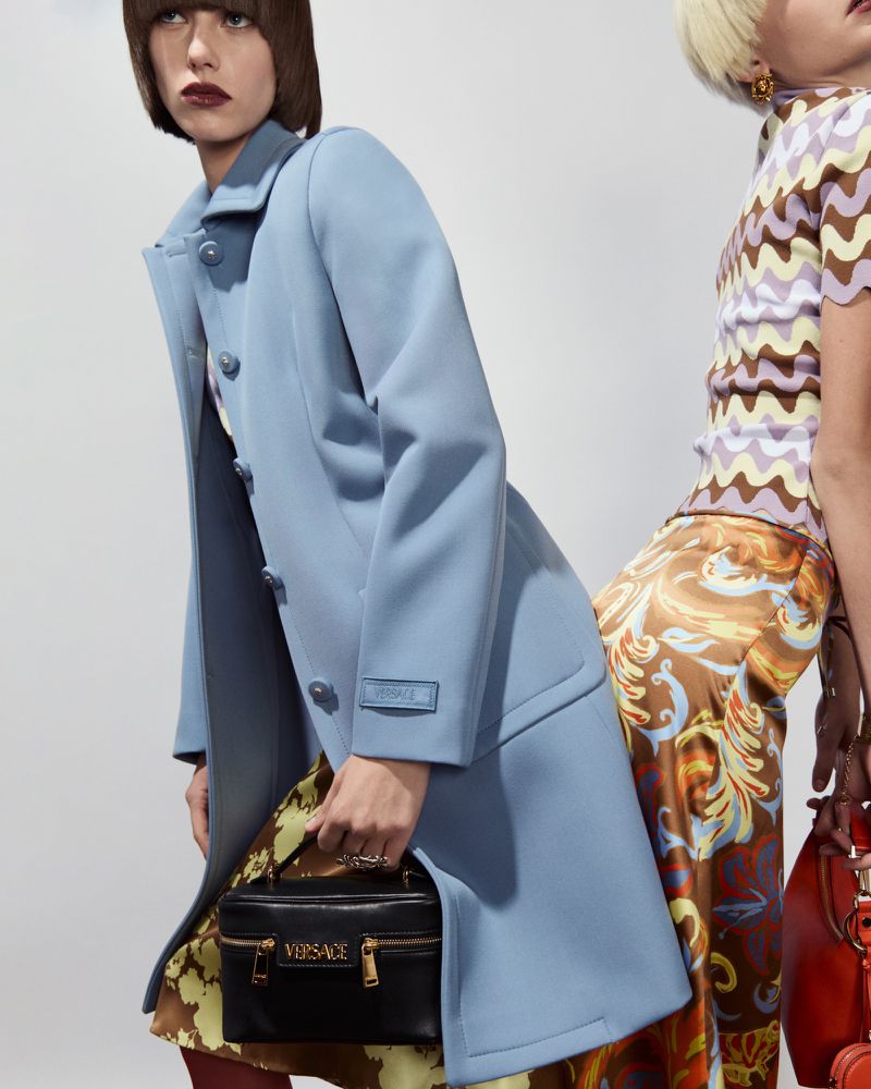 Two fashion models showcasing Versace clothing and accessories against a neutral background, one in a light blue coat and the other in patterned top.