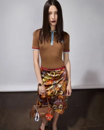 Model wearing a brown polo shirt and patterned skirt holding a brown handbag against a grey backdrop