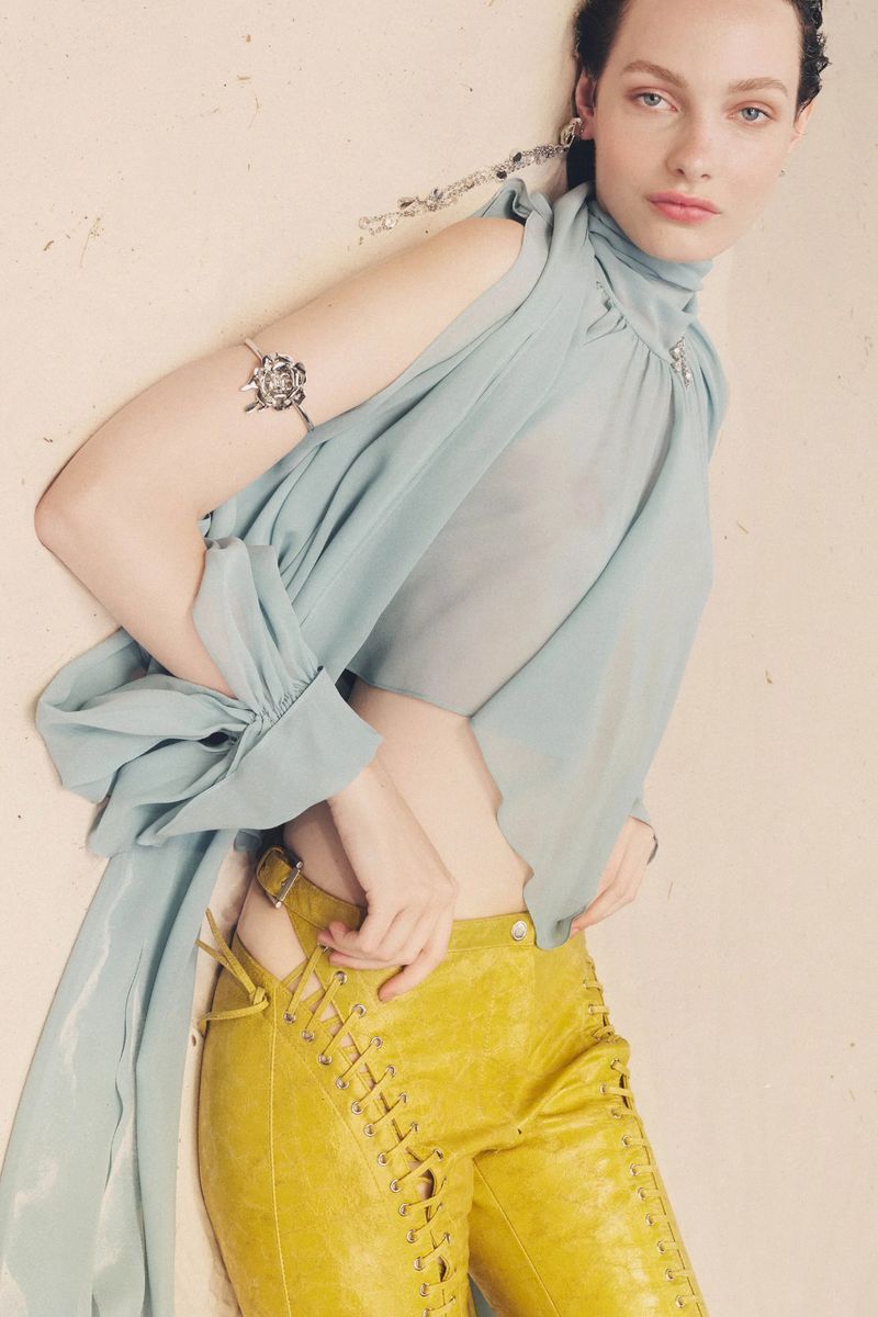 A young woman poses in a light blue chiffon top and yellow leather pants, accessorized with jewelry, against an off-white backdrop.
