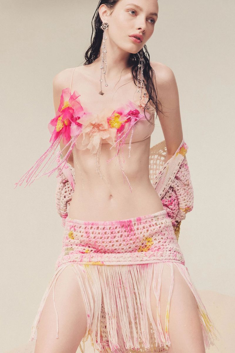 A young woman with long, wet dark hair models a crochet skirt and top with flower accents in a bright, studio-lit fashion shot.