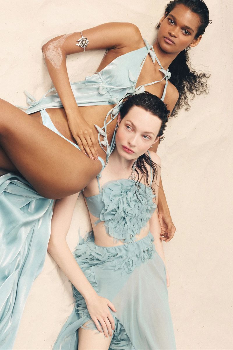 Two women in light blue outfits lying on a neutral surface, gazing at the camera in a fashion editorial style, showcasing makeup and fashion.