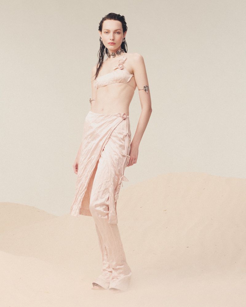 A woman poses in a light pink matching two-piece outfit with a wrap skirt in front of a neutral background, showcasing a fashion-forward aesthetic.