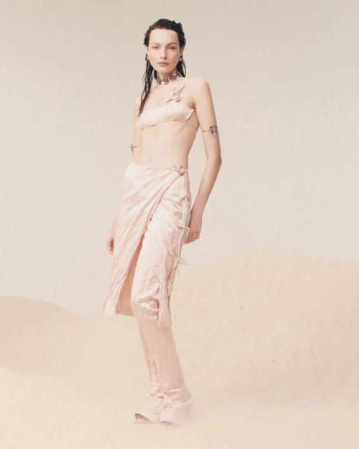 A woman poses in a light pink matching two-piece outfit with a wrap skirt in front of a neutral background, showcasing a fashion-forward aesthetic.