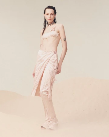 A woman poses in a light pink matching two-piece outfit with a wrap skirt in front of a neutral background, showcasing a fashion-forward aesthetic.