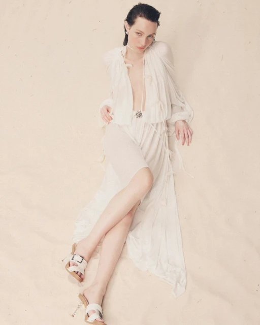 A woman poses in a flowing white outfit and slide sandals on a cream background in a fashion photography shot with a neutral expression.