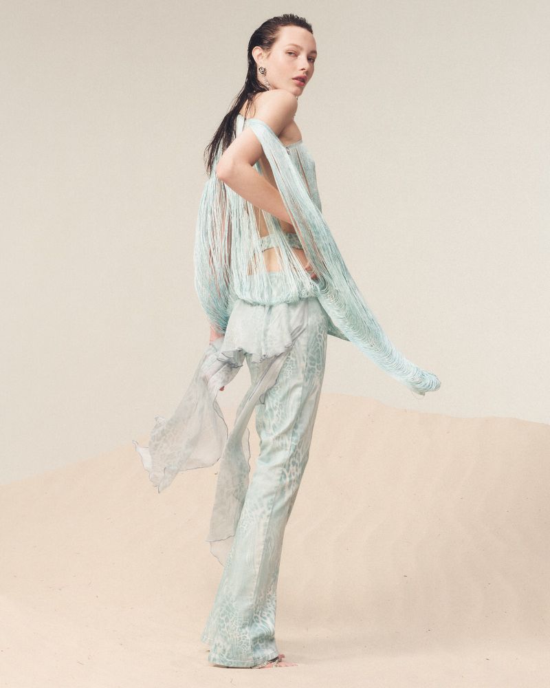 A slender fashion model with dark hair poses in an ethereal aqua, fringed top and flowing pants in a minimal studio setting.