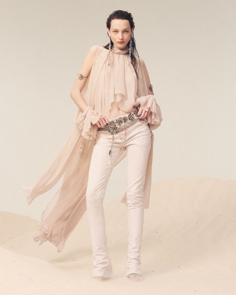 A woman in a flowing peach top and white jeans poses in a studio with a soft sand-like surface, showcasing a fashion-forward look.