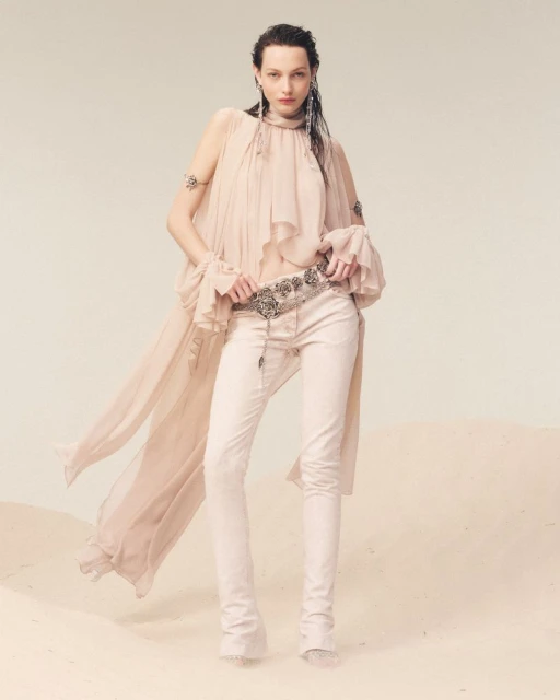 A woman in a flowing peach top and white jeans poses in a studio with a soft sand-like surface, showcasing a fashion-forward look.