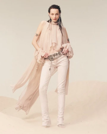 A woman in a flowing peach top and white jeans poses in a studio with a soft sand-like surface, showcasing a fashion-forward look.