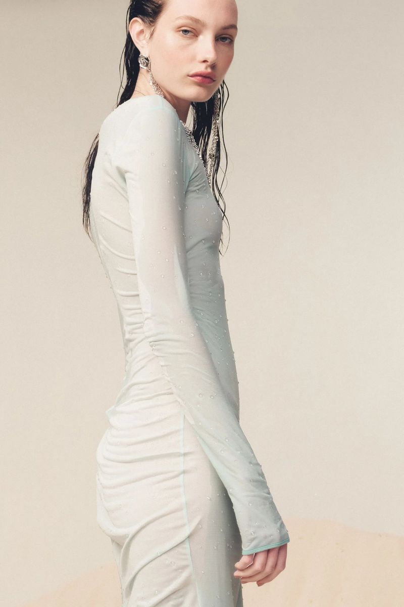 Fashion shot of a slender woman in a light blue, sheer dress with long sleeves and ruched detailing, accessorized with dangling earrings.