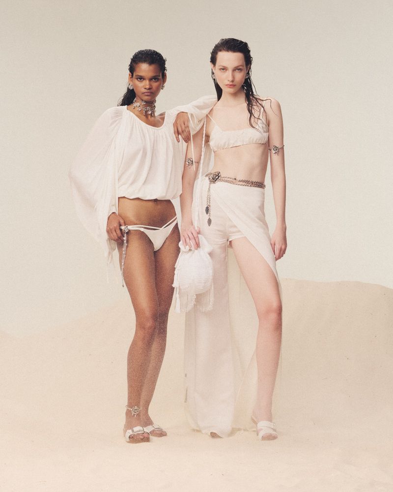 Two female fashion models in light-colored, flowing clothing and jewelry pose in a photo studio with a light tan backdrop.