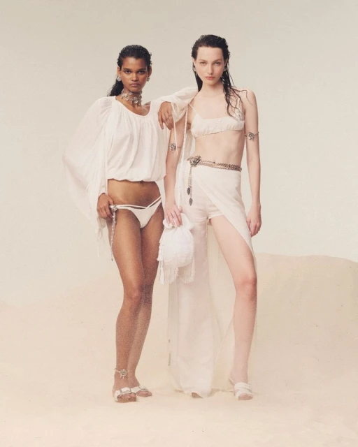 Two female fashion models in light-colored, flowing clothing and jewelry pose in a photo studio with a light tan backdrop.