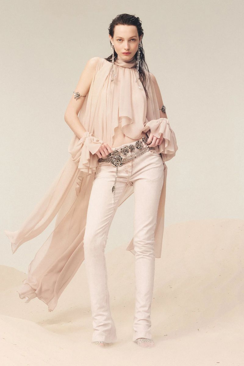 A young adult woman poses in a flowing sleeveless top, jeans, and jewelry in a fashion editorial shot in a bright, neutral studio.