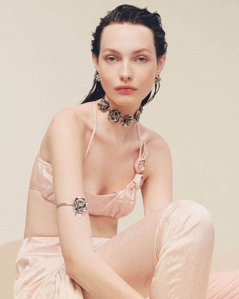 Indoor shot of a woman with dark hair wearing matching light pink satin-like lounge set and jewelry, posing in a soft light environment.