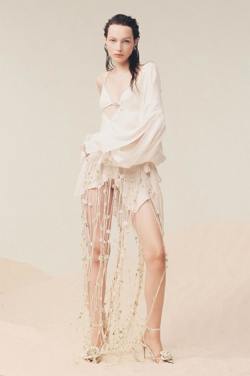 Elegant woman in white ensemble featuring a bra top, draped skirt with macrame detail, and flower-embellished heels poses in studio.