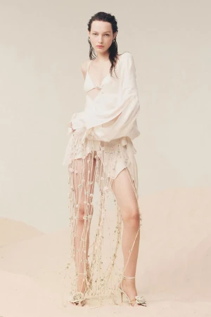 Elegant woman in white ensemble featuring a bra top, draped skirt with macrame detail, and flower-embellished heels poses in studio.