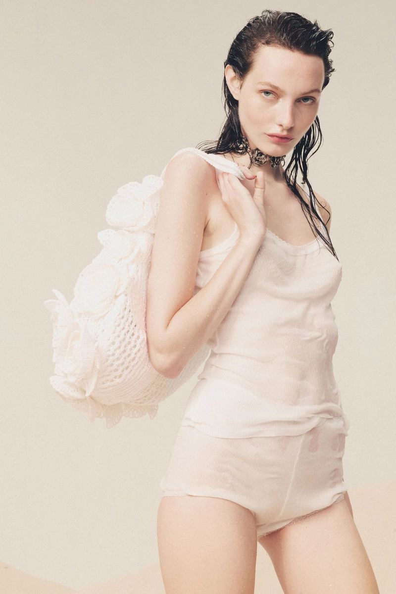 Model with wet hair wearing a white tank top and shorts, holding a floral bag against a neutral background