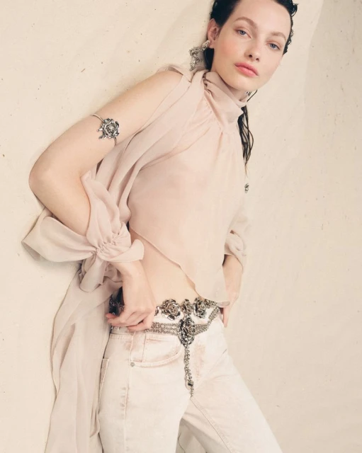 Indoor fashion shot of a young adult woman posing in a light pink sheer top, white pants, and silver jewelry against a beige wall.