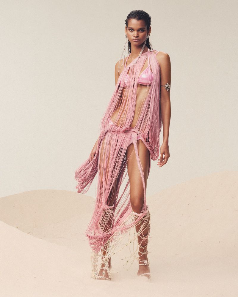Model in a fringed pink outfit and gladiator sandals poses on a sandy surface, against a light beige background in a fashion shot.