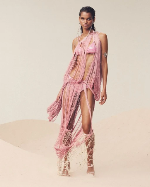 Model in a fringed pink outfit and gladiator sandals poses on a sandy surface, against a light beige background in a fashion shot.