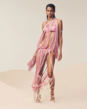 Model in a fringed pink outfit and gladiator sandals poses on a sandy surface, against a light beige background in a fashion shot.