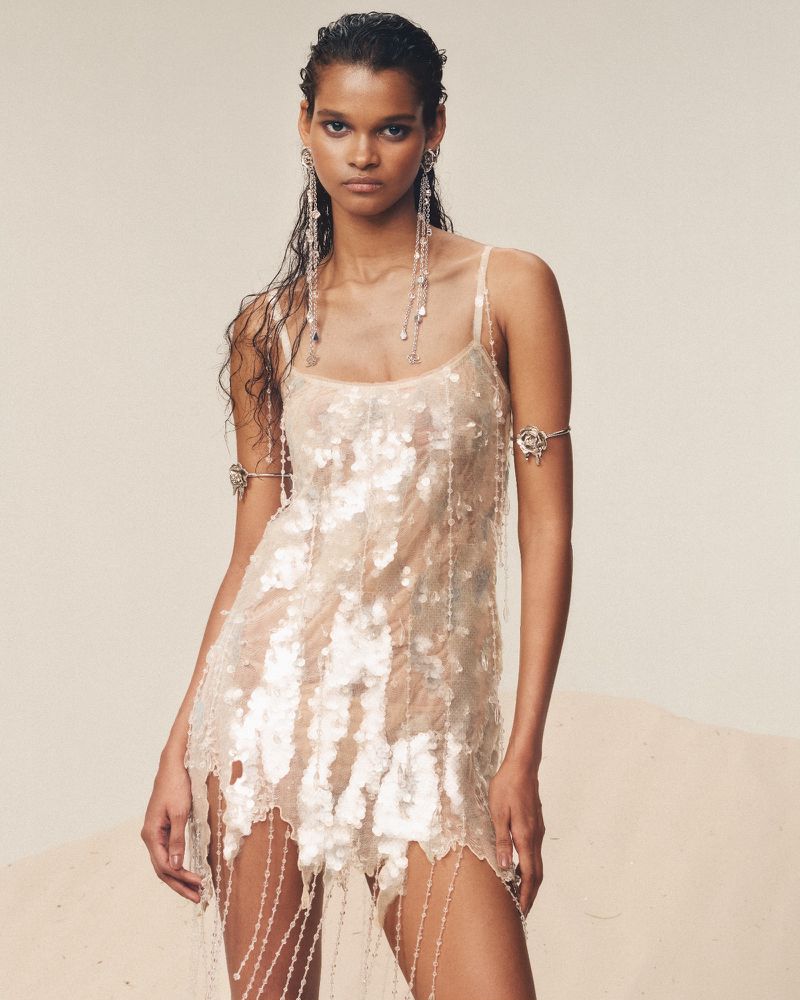 Woman in a shimmering dress with beaded details poses in a studio with wet-look hair, statement earrings, and an arm cuff.