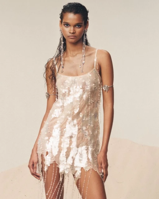 Woman in a shimmering dress with beaded details poses in a studio with wet-look hair, statement earrings, and an arm cuff.
