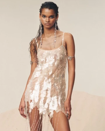 Woman in a shimmering dress with beaded details poses in a studio with wet-look hair, statement earrings, and an arm cuff.