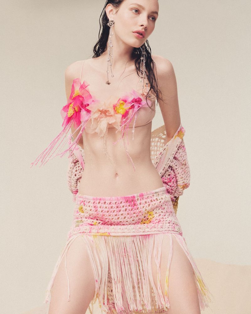 A woman models in a crocheted pink two-piece outfit with floral detailing, styled with delicate silver earrings, against a neutral backdrop.