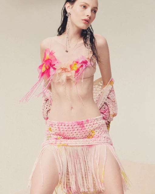 A woman models in a crocheted pink two-piece outfit with floral detailing, styled with delicate silver earrings, against a neutral backdrop.