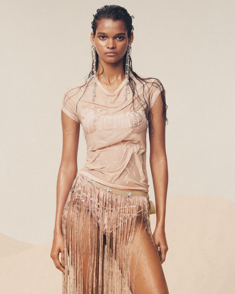 An adult female poses in a wet beige outfit, featuring a T-shirt and fringed skirt, complemented by statement earrings and wet hair.
