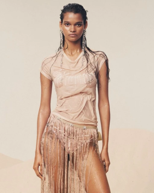 An adult female poses in a wet beige outfit, featuring a T-shirt and fringed skirt, complemented by statement earrings and wet hair.