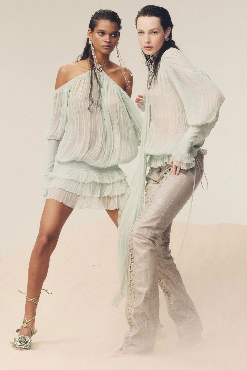 Two fashion models pose in a studio. One wears a light green mini dress with an off-the-shoulder neckline and the other is wearing pants.