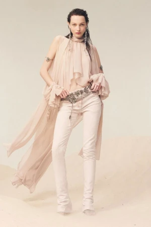 Model wearing a sheer draped blouse and trousers with an embellished belt against a sandy backdrop