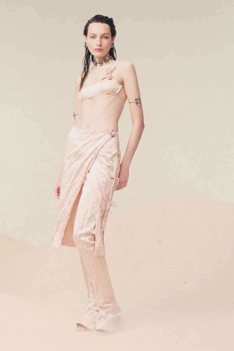 Model in pink satin two-piece outfit standing against a pale neutral sandy backdrop with wet-look dark hair