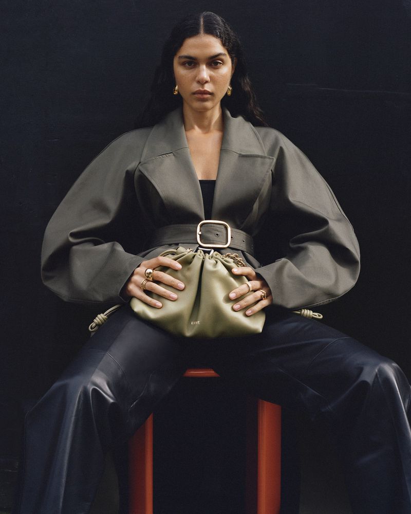 A woman poses while seated on a stool, wearing a gray belted jacket, black leather-like pants, and holding a small green bag.