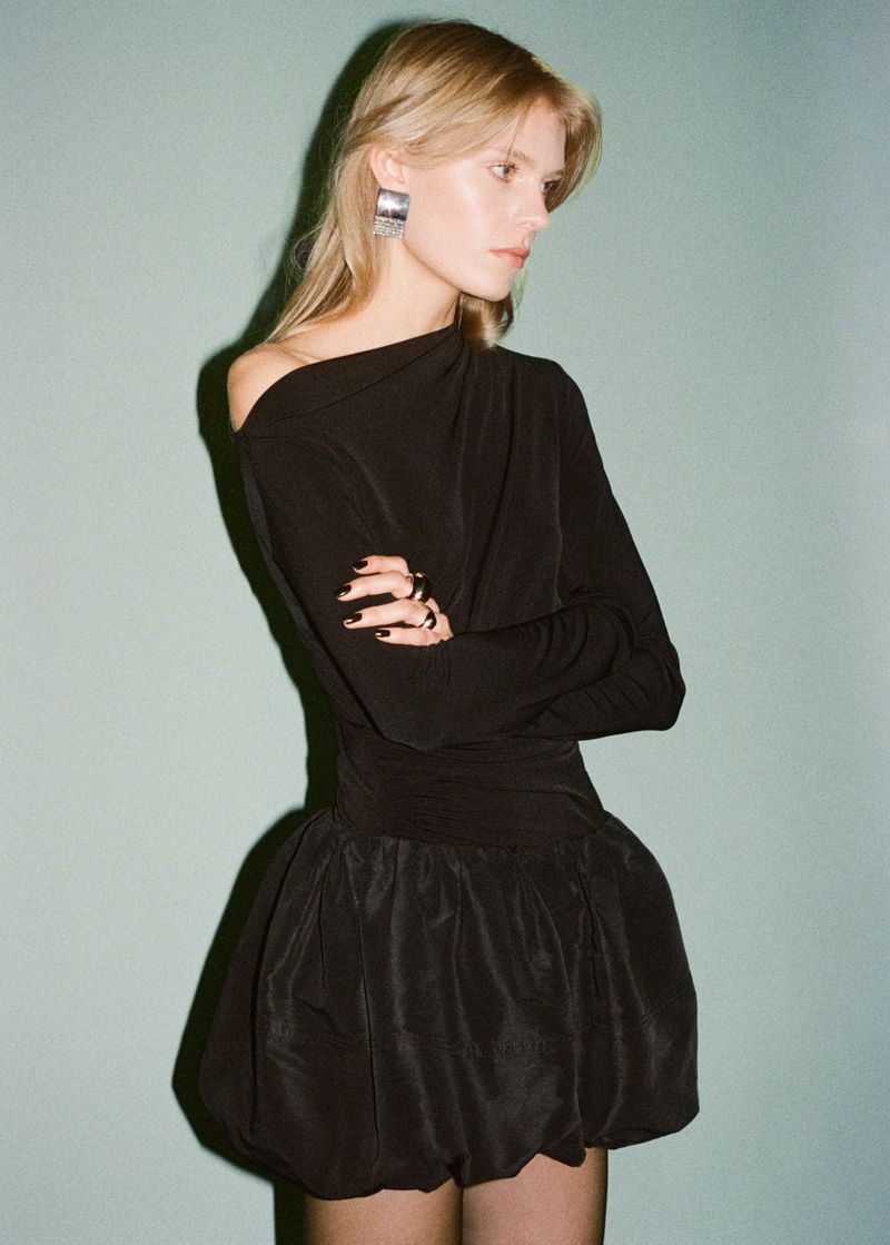 Elegant blonde woman in a black off-the-shoulder dress with statement earrings, posing against a teal backdrop in a fashion-forward studio shot.
