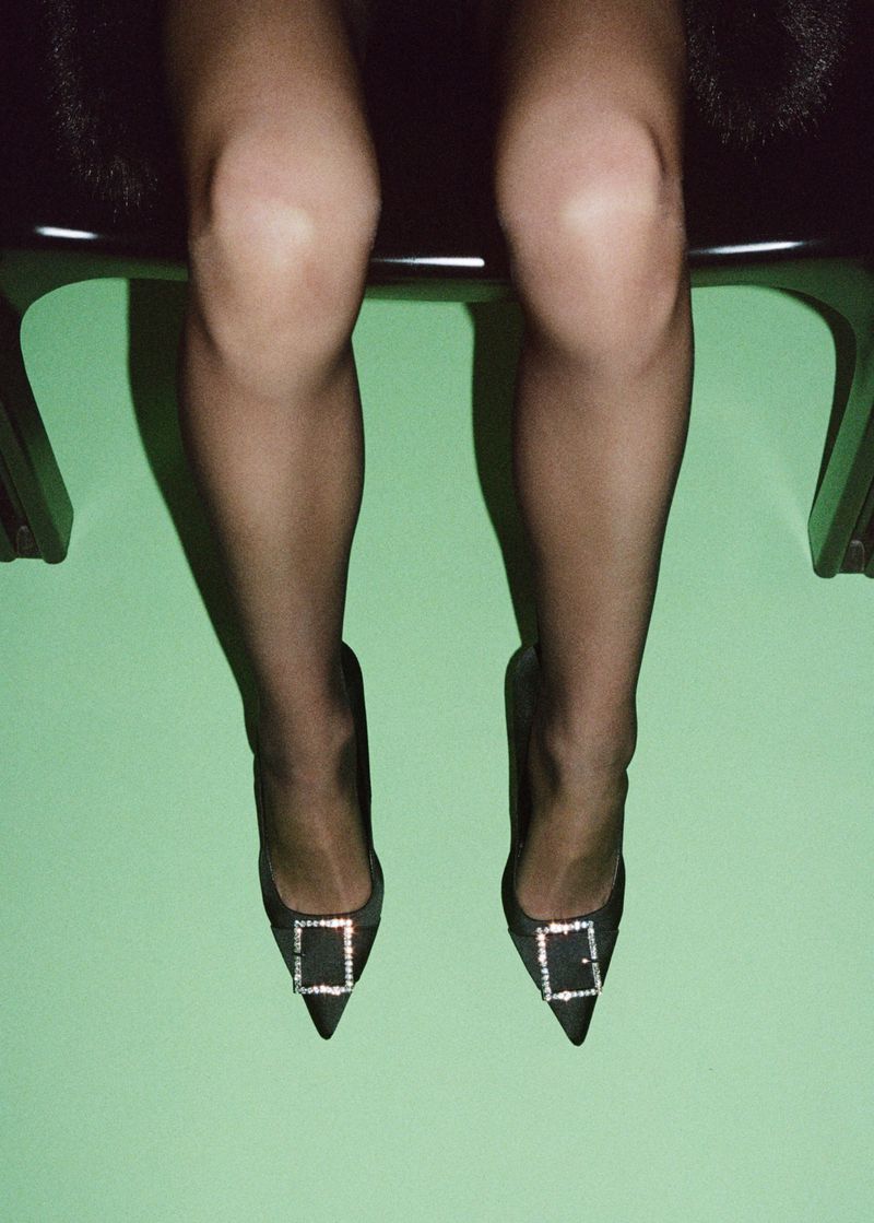 Woman sitting on a chair wearing black pantyhose and black high heels with crystal buckles against a green wall backdrop.