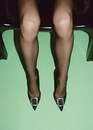 Woman sitting on a chair wearing black pantyhose and black high heels with crystal buckles against a green wall backdrop.