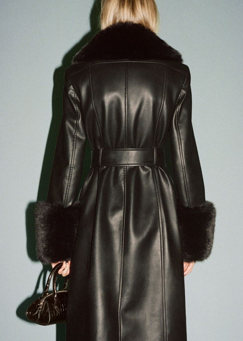 Rear shot of a woman in a belted black leather coat with fur trim and a matching handbag against a plain blue-grey background.