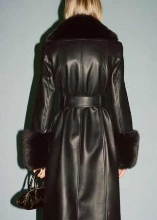 Rear shot of a woman in a belted black leather coat with fur trim and a matching handbag against a plain blue-grey background.