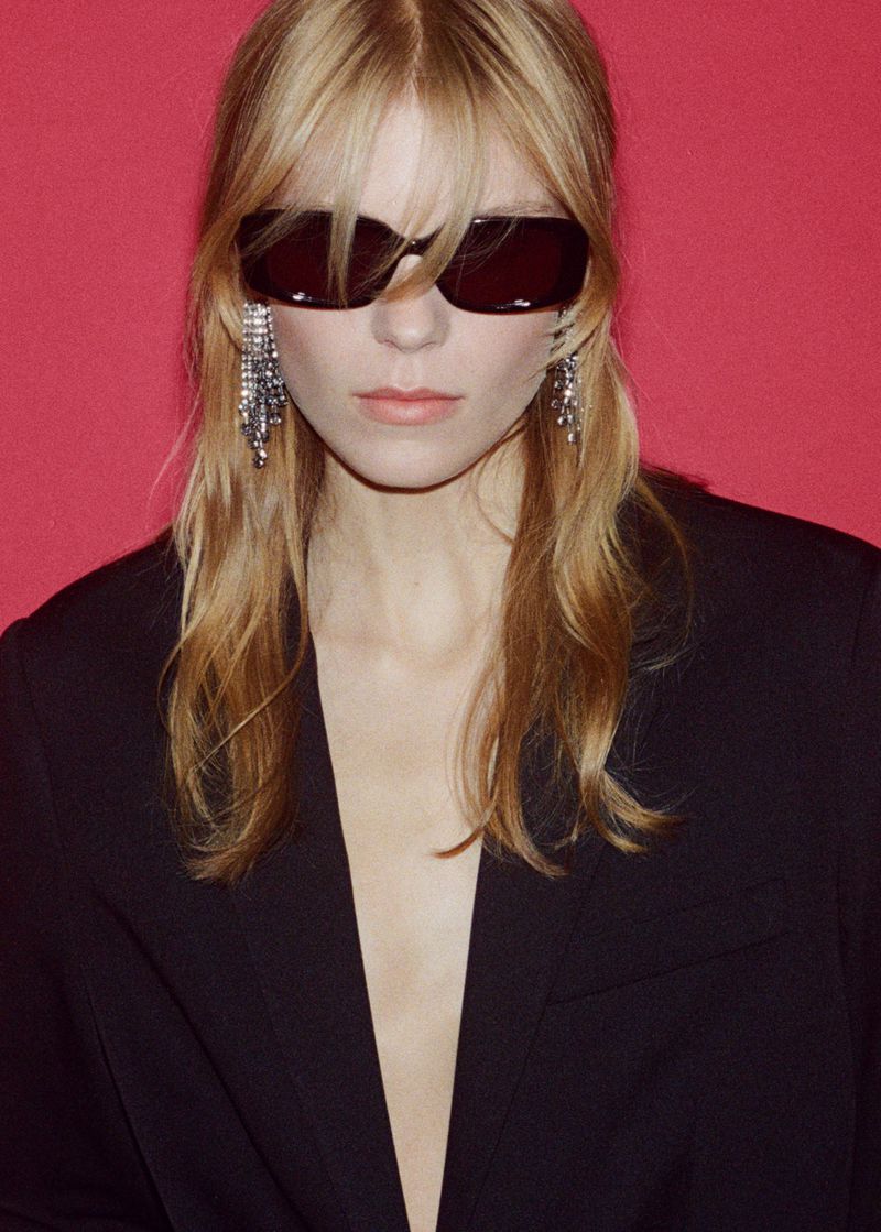 Model wearing dark sunglasses, crystal fringe earrings, and a black jacket against a solid red background